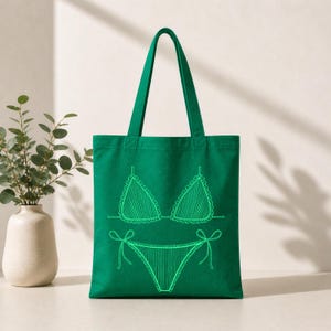 May include: A green tote bag featuring a neon green outline of a bikini. The bikini top has a scalloped edge and the bottoms have tie details. The bag has two green handles and is made of a sturdy fabric, perfect for carrying beach essentials.