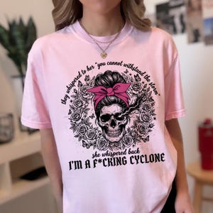 Può includere: Una t-shirt rosa chiaro con una grafica nera di un teschio con uno chignon e un fiocco rosa, circondato da rose e farfalle. Il testo recita: "they whispered to her you cannot withstand the storm she whispered back I'M A F*CKING CYCLONE."