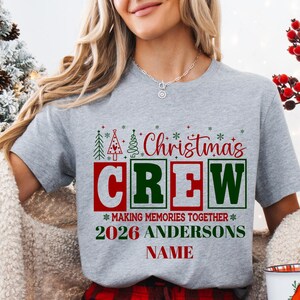 Pu&ograve; includere: Una t-shirt grigio melange con il testo rosso e verde "Christmas CREW" sopra "MAKING MEMORIES TOGETHER", "2026 ANDERSONS" e "NAME". Il design include piccole icone di alberi di Natale verdi e rossi e accenti a fiocco di neve.