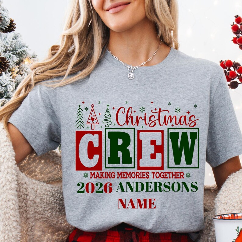 Pu&ograve; includere: Una t-shirt grigio melange con il testo rosso e verde "Christmas CREW" sopra "MAKING MEMORIES TOGETHER", "2026 ANDERSONS" e "NAME". Il design include piccole icone di alberi di Natale verdi e rossi e accenti a fiocco di neve.