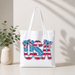 Op de afbeelding: Witte draagtas met het woord "USA" in rode, witte en blauwe letters in spijkerbroekstijl. De letters zijn versierd met strikken en sterren, die doen denken aan de Amerikaanse vlag.