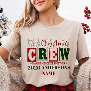 Pu&ograve; includere: Una t-shirt beige a maniche corte con una grafica che riporta il testo "Christmas CREW MAKING MEMORIES TOGETHER 2026 ANDERSONS NAME" in rosso e verde. Il design include piccole icone di alberi di Natale e accenti a forma di fiocco di neve.