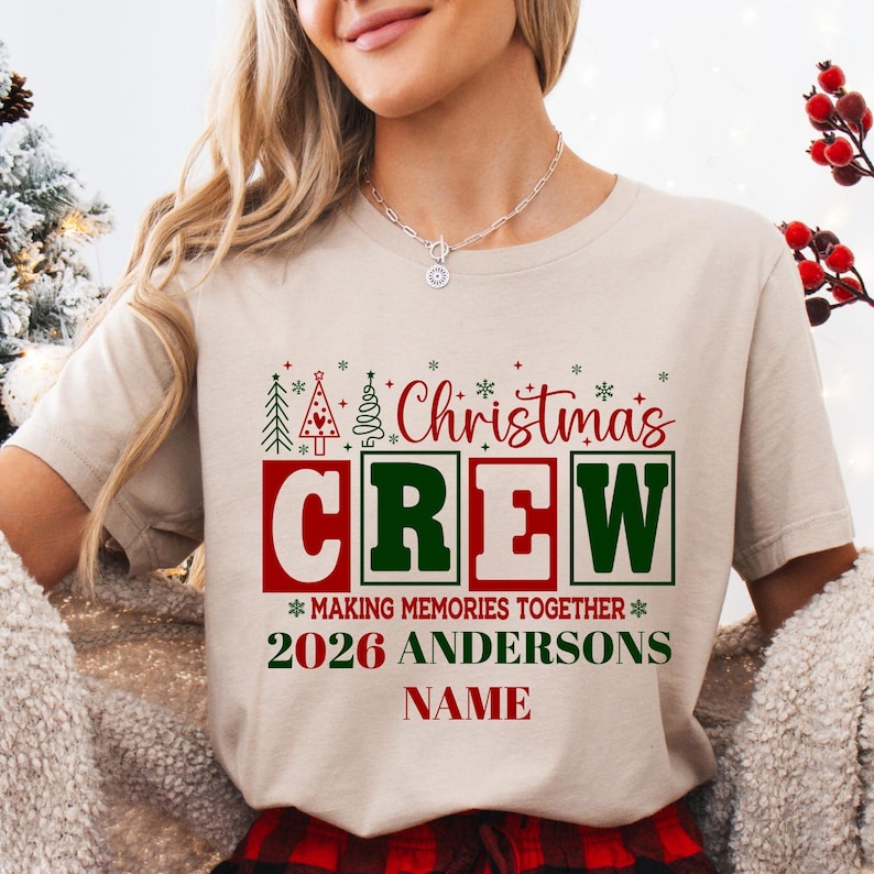 Pu&ograve; includere: Una t-shirt beige a maniche corte con una grafica che riporta il testo "Christmas CREW MAKING MEMORIES TOGETHER 2026 ANDERSONS NAME" in rosso e verde. Il design include piccole icone di alberi di Natale e accenti a forma di fiocco di neve.