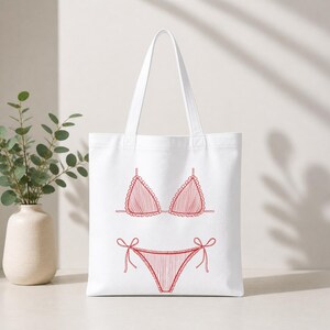 May include: White tote bag featuring a red line drawing of a bikini. The bikini top has a triangle shape with ruffled edges, and the bottom has tie sides. The bag has a simple design, perfect for carrying beach essentials.