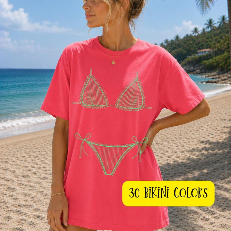 May include: A coral-colored t-shirt featuring a green bikini outline. The design includes a bra and bikini bottoms with tie details. The text "30 BIKINI COLORS" is displayed in a yellow rectangle at the bottom of the shirt. The shirt is worn on a beach.