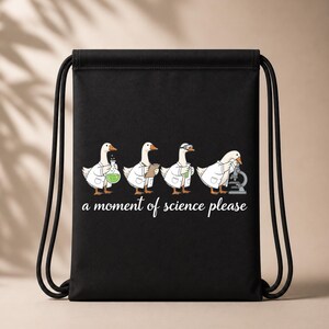 May include: A black drawstring backpack featuring a graphic of four white geese wearing lab coats. The geese are performing scientific tasks with a flask, clipboard, test tube, and microscope. Below the geese is the text, a moment of science please.