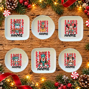 May include: Six white ceramic plates featuring colourful Christmas-themed illustrations of cartoon characters wearing festive hats. Each plate displays a unique character graphic above the black text NAME, set against a red and white striped background.