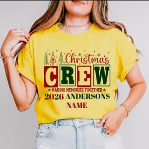 Pu&ograve; includere: Una t-shirt gialla a maniche corte con una grafica festiva che riporta il testo Christmas CREW, MAKING MEMORIES TOGETHER, 2026 ANDERSONS e NAME. Il design include scritte rosse e verdi, alberi di Natale stilizzati e fiocchi di neve.