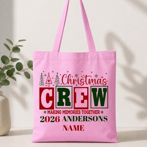 Pu&ograve; includere: Una borsa tote rosa chiaro con una grafica festiva che include il testo rosso Christmas, il testo verde e rosa CREW e la frase MAKING MEMORIES TOGETHER 2026 ANDERSONS NAME in rosso e verde, accentuata da piccole icone di alberi di Natale.