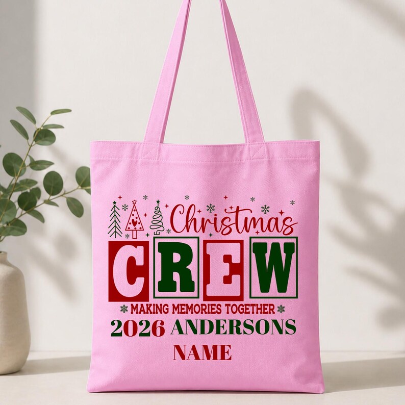 Pu&ograve; includere: Una borsa tote rosa chiaro con una grafica festiva che include il testo rosso Christmas, il testo verde e rosa CREW e la frase MAKING MEMORIES TOGETHER 2026 ANDERSONS NAME in rosso e verde, accentuata da piccole icone di alberi di Natale.