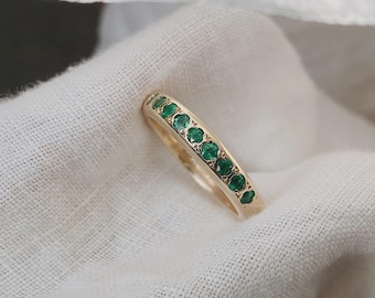 Puede incluir: Un anillo de banda de oro con una hilera de pequeñas piedras preciosas redondas de color verde vibrante engastadas a lo largo de la superficie superior. El metal tiene un acabado pulido y las piedras están dispuestas en línea.