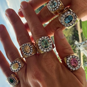 Puede incluir: Una colección de siete anillos de oro, cada uno con una banda ancha y una piedra preciosa ovalada central rodeada por un halo de pequeñas piedras blancas redondas. Las gemas incluyen azul claro, azul oscuro, verde, amarillo, rosa, ópalo y verde oscuro.