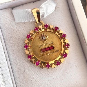 Puede incluir: Un colgante redondo de oro con una cruz central con un pequeño diamante, el texto "QU'HIER" y "QUE DEMAIN" separados por una barra rectangular de tres rubíes rojos, rodeado por un borde de pequeños corazones de oro y rubíes alternados.