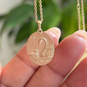 Può includere: Una collana con ciondolo ovale in oro caratterizzata da un design in rilievo di un cigno e un cignetto che nuotano sull'acqua. Il ciondolo ha una finitura opaca testurizzata e include una piccola gemma trasparente a taglio rotondo in alto.