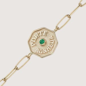 May include: A gold chain bracelet featuring an octagonal pendant with a textured surface. The pendant is embossed with the text PORTE BONHEUR and has a small, round, bezel-set green gemstone centered in the middle of the design.