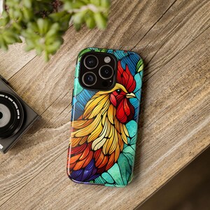 Puede incluir: Una funda de teléfono colorida con un diseño de gallo estilizado. El gallo tiene una cresta roja, plumas amarillas y naranjas, y un fondo azul y verde. La funda está sobre una superficie de madera.
