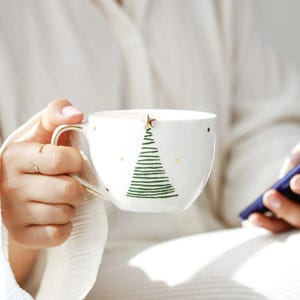 May include: White ceramic mug with a green Christmas tree design and gold accents. The mug features a gold star on top of the tree and gold dots around the rim. The handle is also gold.
