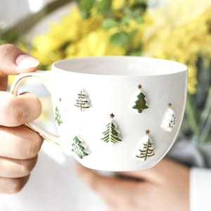 Puede incluir: Taza de cerámica blanca con borde dorado, adornada con diseños en relieve de árboles de Navidad verdes y blancos y pequeñas estrellas doradas. La taza tiene un asa curva y una abertura ancha.