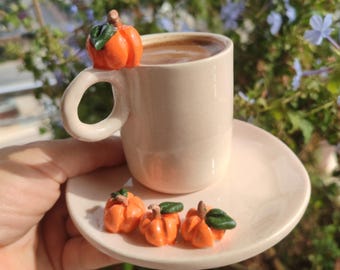 May include: A small, light pink ceramic coffee cup and saucer set. The cup is filled with coffee and adorned with a miniature orange pumpkin on the rim. Three additional pumpkin decorations sit on the saucer. The background is blurred with greenery and blue flowers.