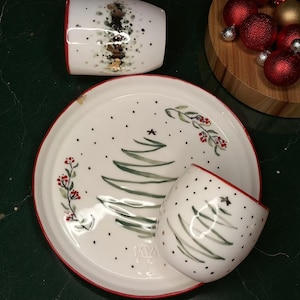 Puede incluir: Plato y tazas navideños de cerámica blanca con bordes rojos. El plato presenta un diseño de árbol de Navidad verde con bayas rojas y una estrella. Las tazas tienen un diseño similar y lunares negros.