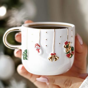 Puede incluir: Taza navideña de cerámica blanca con detalles dorados y asa dorada. La taza está decorada con adornos colgantes, como un muñeco de nieve, campanas y un árbol de Navidad. La taza está llena de un líquido oscuro.