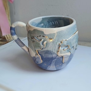 May include: A ceramic mug with a blue and white watercolor design and gold accents. The mug features a map outline and the text "JAYA STUDIO". The handle is striped blue and white.