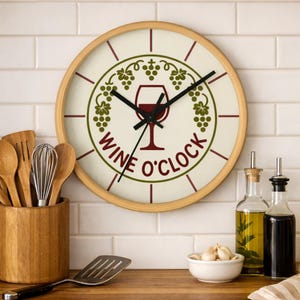 Może przedstawiać: Okrągły zegar ścienny z jasną drewnianą ramą. Tarcza zegara jest kremowa z grafiką kieliszka do wina i napisem "WINE O'CLOCK". Zielone wzory winogron i liści otaczają kieliszek do wina. Dekoracja kuchni.
