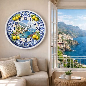 Puede incluir: Un reloj de pared redondo con marco blanco y diseño de mosaico azul y blanco. La esfera del reloj presenta una escena costera con limones, un pueblo y veleros. Las manecillas del reloj son blancas.