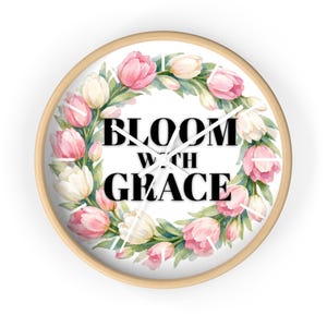 May include: A round wall clock with a light wood frame. The clock face features a wreath of pink and white tulips surrounding the words "BLOOM WITH GRACE" in black. The clock hands are white.