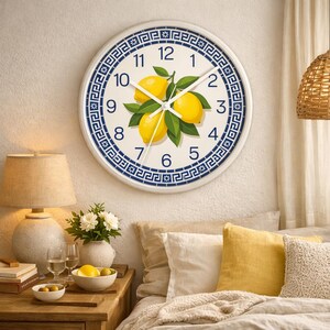 Può includere: Un orologio da parete rotondo bianco con un bordo a motivi blu e bianchi. Il quadrante dell'orologio presenta un design a limone e foglie. Le lancette dell'orologio sono nere. L'orologio è appeso a una parete bianca.