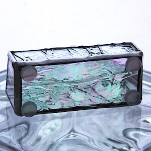 Peut inclure: Une petite bo&icirc;te rectangulaire en verre avec un cadre noir et un verre iridescent bleu, vert et violet. La bo&icirc;te a quatre pieds en caoutchouc sur le fond.