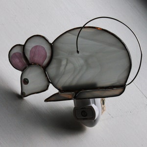 May include: A stained glass nightlight shaped like a gray mouse with pink ears. The nightlight is plugged into an outlet.