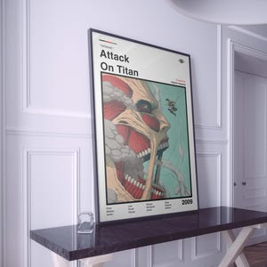 Op de afbeelding: Een ingelijste poster met de titel "Attack On Titan" en een grafische illustratie van een titanengezicht. Het kunstwerk gebruikt rode, beige en groene tinten. De poster wordt op een donkere tafel getoond.