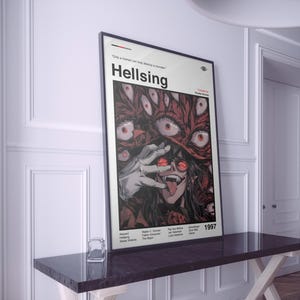 Peut inclure: Affiche encadrée avec le titre "Hellsing" et une illustration graphique d'un personnage aux yeux rouges et un chapeau rouge. L'affiche comprend l'année 1997 et une liste de noms. L'affiche est exposée sur une table sombre.