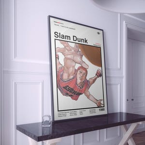 Puede incluir: Un póster enmarcado con un borde negro, que representa a un jugador de baloncesto con un uniforme rojo, con el título "Slam Dunk". El póster se muestra sobre una mesa oscura. El póster también incluye el año 1990.