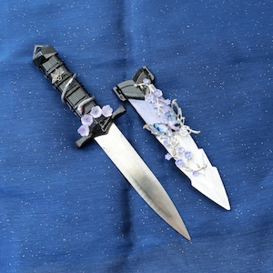 Op de afbeelding: Een decoratieve dolk met een zilveren lemmet en een zwarte handgreep gewikkeld in een textuur. De handgreep is versierd met een zilveren slang en paarse bloemaccenten. De schede is wit met paarse bloemen en vlinders.