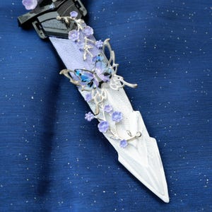 Op de afbeelding: Een decoratieve dolk met een wit lemmet en een zwart handvat. De dolk is versierd met zilverkleurige metalen vlinders en paarse bloemaccenten. De vlinders hebben blauwe en paarse vleugels. De achtergrond is een donkerblauwe stof met kleine witte stippen.
