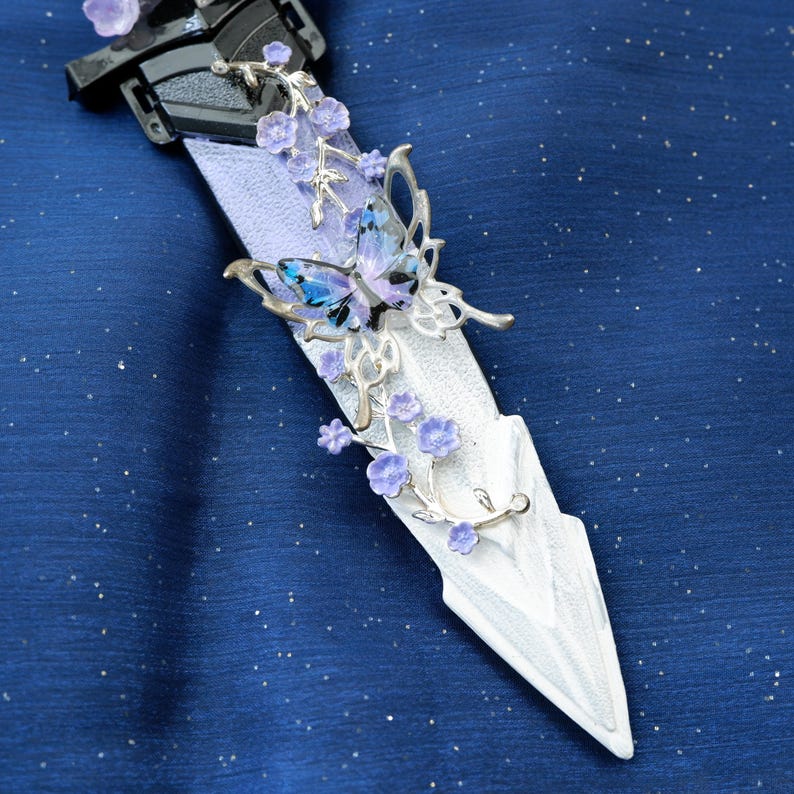 Op de afbeelding: Een decoratieve dolk met een wit lemmet en een zwart handvat. De dolk is versierd met zilverkleurige metalen vlinders en paarse bloemaccenten. De vlinders hebben blauwe en paarse vleugels. De achtergrond is een donkerblauwe stof met kleine witte stippen.