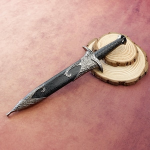 Può includere: Un pugnale decorativo con manico e fodero neri, con dettagli ornamentali in metallo color argento. Il pugnale è appoggiato su una fetta di legno su uno sfondo marrone rosato. La lama non è visibile.