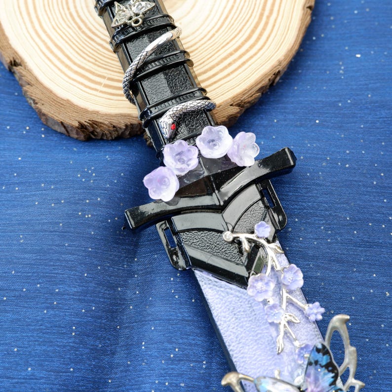 Op de afbeelding: Een decoratief fantasiezwaard met een zwarte handgreep en bescherming, versierd met een zilveren slang, paarse bloemen en een vlinder. Het lemmet is lichtpaars. Het zwaard ligt op een blauw oppervlak.