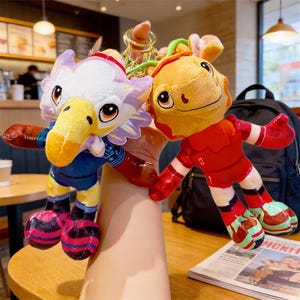 May include: Two plush keychains: one eagle-like with a white head, purple accents, and a yellow beak, wearing a blue shirt. The other is a yellow creature in a red outfit. Both have large eyes and are held together by a gold keychain.