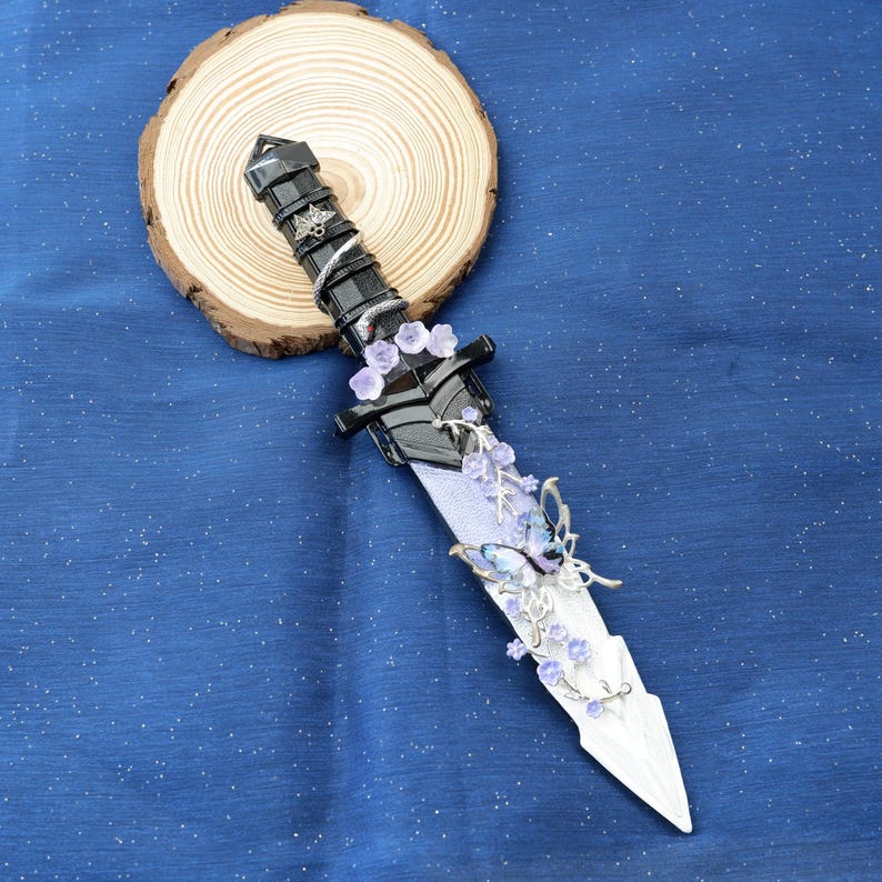Op de afbeelding: Een decoratieve dolk met een zwarte handgreep en een zilveren lemmet. De handgreep is versierd met zilveren accenten en een spiraalvormig ontwerp. Het lemmet heeft een bloemen- en vlinderontwerp met paarse en blauwe accenten. De dolk wordt getoond op een houten schijf.