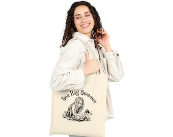 May include: A cream-colored canvas tote bag featuring a black graphic illustration of a figure emerging from water with the text "Sea Hag Summer" arched above the image. The bag has two shoulder straps and a rectangular shape for carrying items.