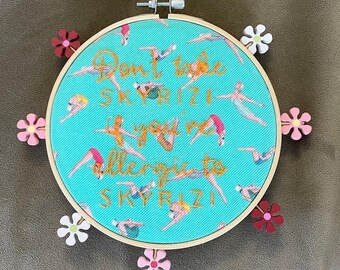 May include: A circular wooden embroidery hoop containing turquoise fabric with a repeating pattern of swimmers. Gold embroidered text reads: Don't take SKYRIZI if you're allergic to SKYRIZI. Six small wooden flower shapes in pink, red, and white are attached.