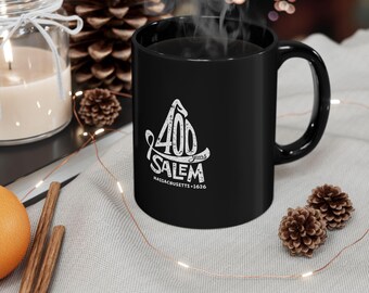 May include: A black ceramic mug featuring a white graphic design with the text 400 years SALEM MASSACHUSETTS 1626. The design includes a stylized witch hat silhouette. The mug has a smooth finish, a curved handle, and is filled with a hot beverage.