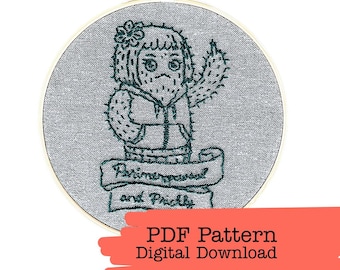 May include: An embroidery hoop with a gray fabric background features a cactus person design in green thread. The cactus character is wearing a hoodie and has a flower on its head. Below the character is a banner that reads "Prim and Prickly".