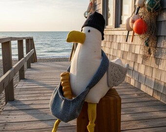 May include: A plush seagull toy with a white body, yellow beak, and yellow webbed feet. The seagull wears a black knit beanie and a grey scarf, and carries a grey fabric shoulder bag containing a yellow baguette-shaped item.