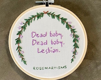 May include: A circular wooden embroidery hoop containing white fabric with hand-stitched purple text that reads "Dead baby. Dead baby. Lesbian." and "ROSEMARYISMS" at the bottom, framed by an embroidered green and purple floral wreath.