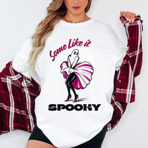 Op de afbeelding: Wit lange mouwen shirt met de tekst "Some Like it Spooky". De afbeelding toont een geest in een jurk en hoge hakken. Het shirt wordt gecombineerd met een rood-zwart geruit overhemd.