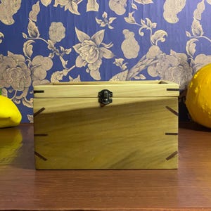 May include: A rectangular wooden box with a metal clasp, light brown in colour with dark brown detailing. The box is on a wooden surface, with a yellow pumpkin and squash in the background.
